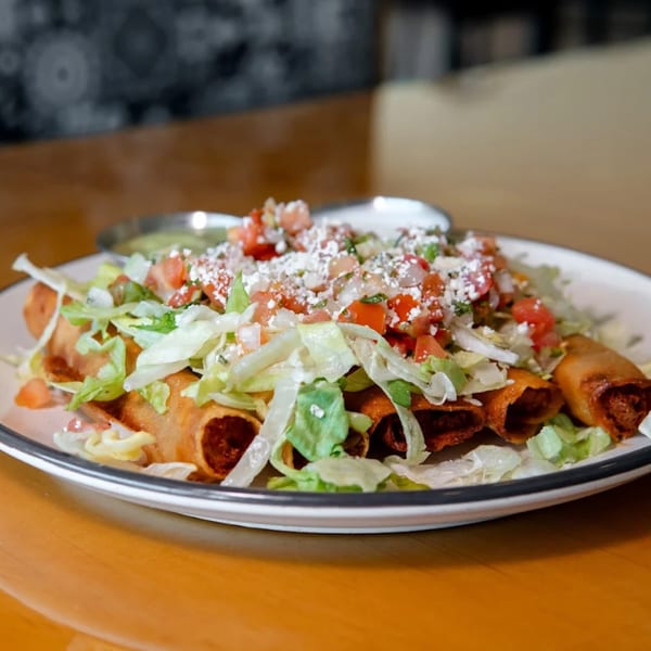 Crispy taquitos topped with shredded lettuce, diced tomatoes, crumbled cheese, and a side of salsa.