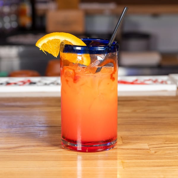 A vibrant orange and red cocktail with an orange slice and straw sits on a wooden bar.