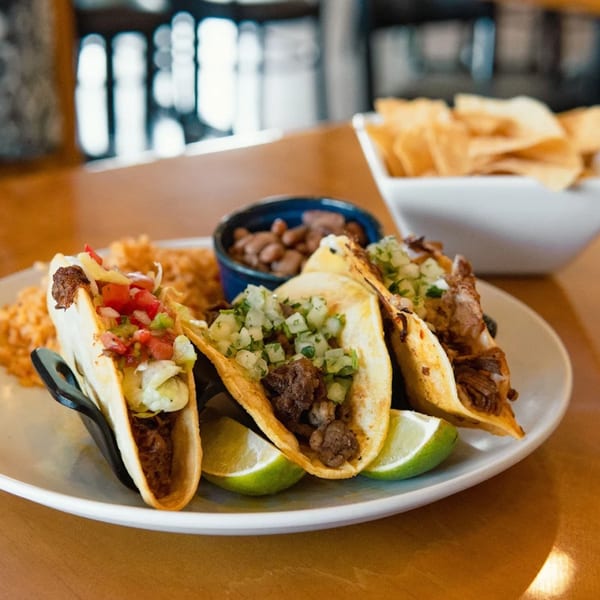 Three Jefe's Cantina + Cocina tacos with rice, beans, and tortilla chips on a wooden table.